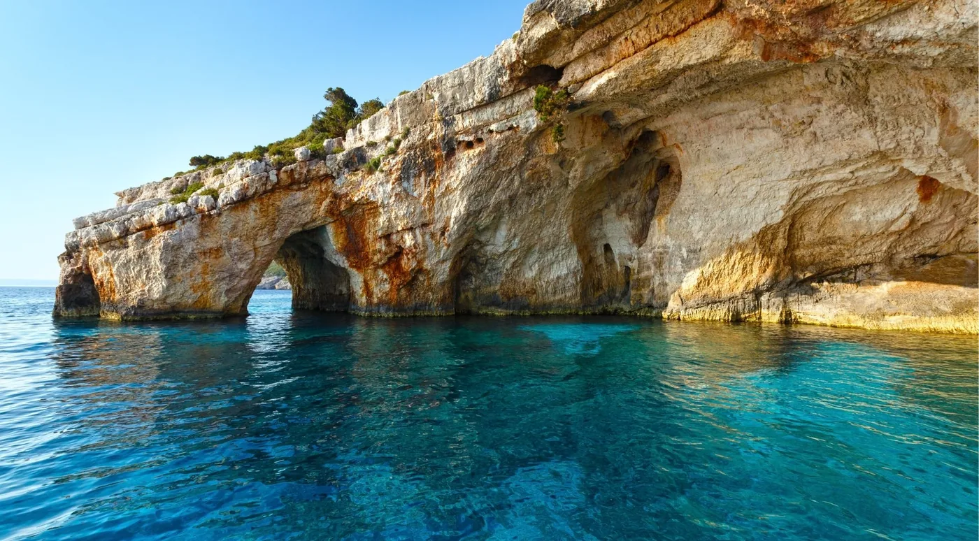 A low sea arch cut into a limestone headland on the Zakynthos coast, with clear turquoise water in the foreground