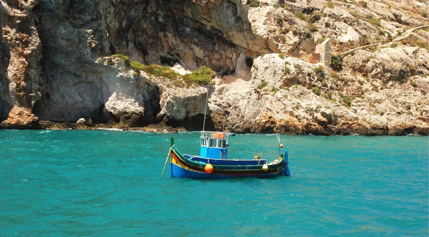 A small painted fishing boat anchored below eroded ochre and grey limestone cliffs