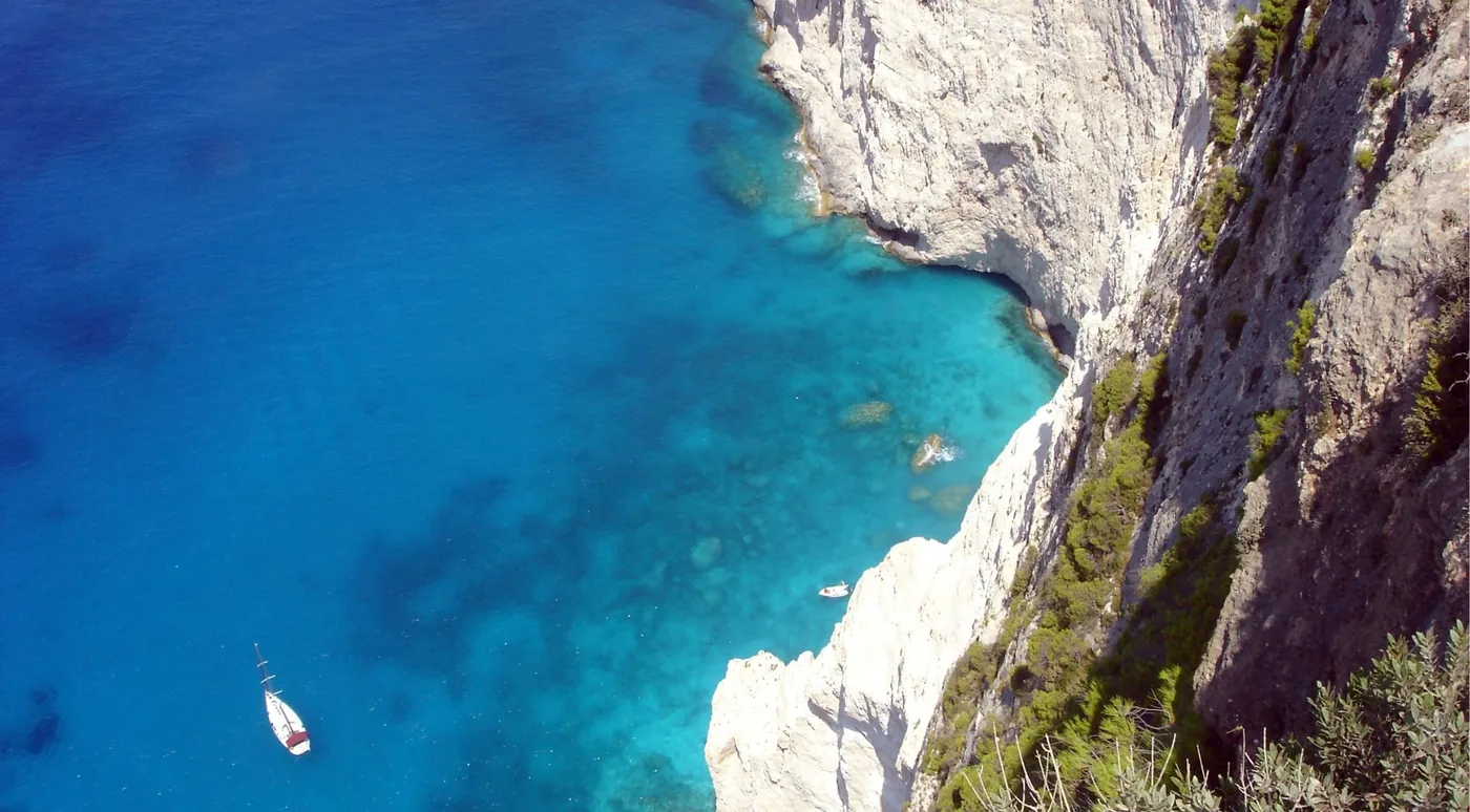 Looking down a sheer white cliff face to a sailing boat and a small motorboat on deep blue water